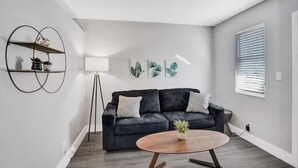 Una sala de estar moderna con un sofá gris oscuro, una mesa de centro redonda de madera y una lámpara de pie. En la pared hay tres grabados botánicos enmarcados y una estantería metálica negra con objetos decorativos.