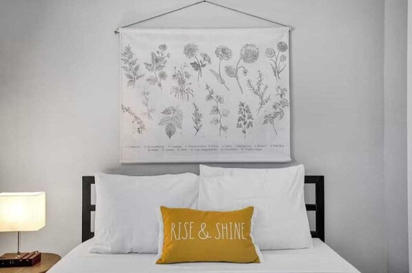 Un letto rifatto con lenzuola bianche e un cuscino giallo senape con la scritta “RISE & SHINE”. Sopra il letto, un'illustrazione botanica appesa al muro.