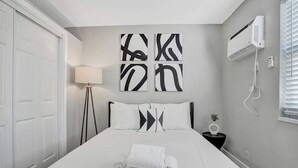 A neatly made bed with a rolled white towel, a bedside lamp, and a wall-mounted air conditioner.