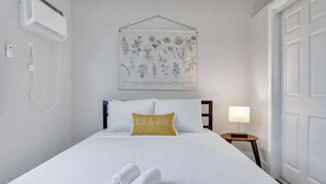 A hotel room with a bed, a wall-mounted air conditioner, a bedside table with a lamp, and a framed botanical print above the bed. - Urban Escape 1BR Near Las Olas (Fort Lauderdale)