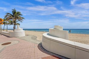 Une promenade en bord de mer, avec une structure blanche incurvée, des palmiers et un panneau indiquant un passage piéton.