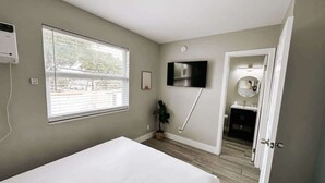 Un dormitorio con cama, televisor montado en la pared, ventana y puerta que da al baño.