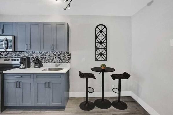 A modern kitchen with grey cabinets, a patterned backsplash, a coffee maker, and a small table with stools.