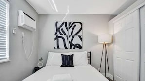 Quarto de hotel moderno com cama, ar condicionado fixado na parede, abajur e uma grande obra de arte abstrata.