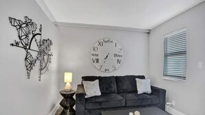 Uma sala de estar moderna com um sofá cinza escuro, um relógio de parede redondo, um objeto decorativo na parede, uma mesinha com um abajur e uma janela com persianas.