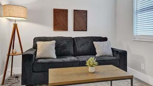 Un salón con un sofá de color gris oscuro, dos adornos en la pared, una lámpara de pie y una mesa de centro con una planta.
