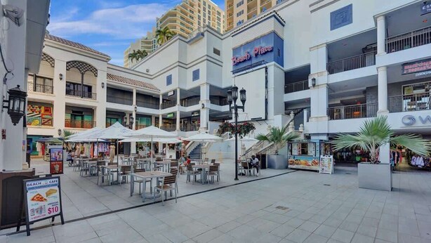 A spacious outdoor area with multiple levels of shops and restaurants, including a visible sign for 'Beach Place' and a 'Beach Cafe' offering pizza.