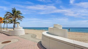 Een boulevard aan het strand met een gebogen witte constructie, palmbomen en een voetgangersoversteekbord.