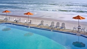 Una piscina con agua cristalina azul, rodeada de sillas de descanso y sombrillas naranjas, con una playa y el océano al fondo.