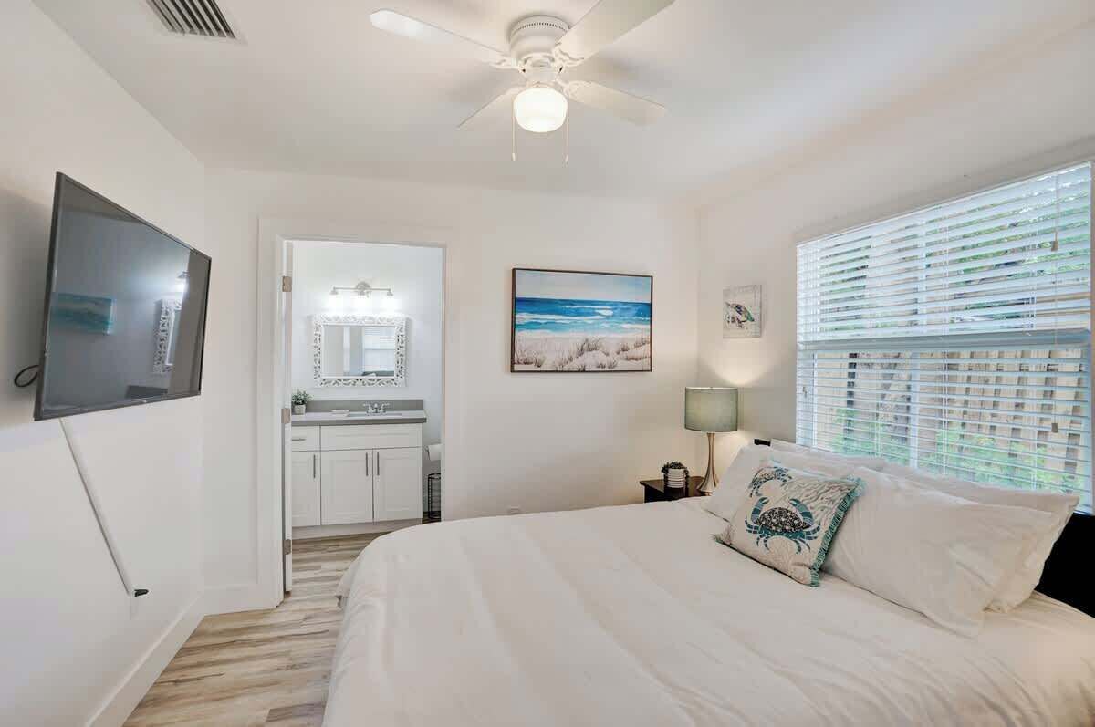 A bedroom with a bed, a ceiling fan, a TV, a bathroom with a sink and mirror, and a window with blinds.