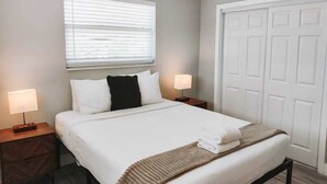 Un lit bien fait, avec du linge de lit blanc, un oreiller noir et un plaid beige. Il y a deux tables de chevet avec des lampes, et une porte blanche avec des panneaux.