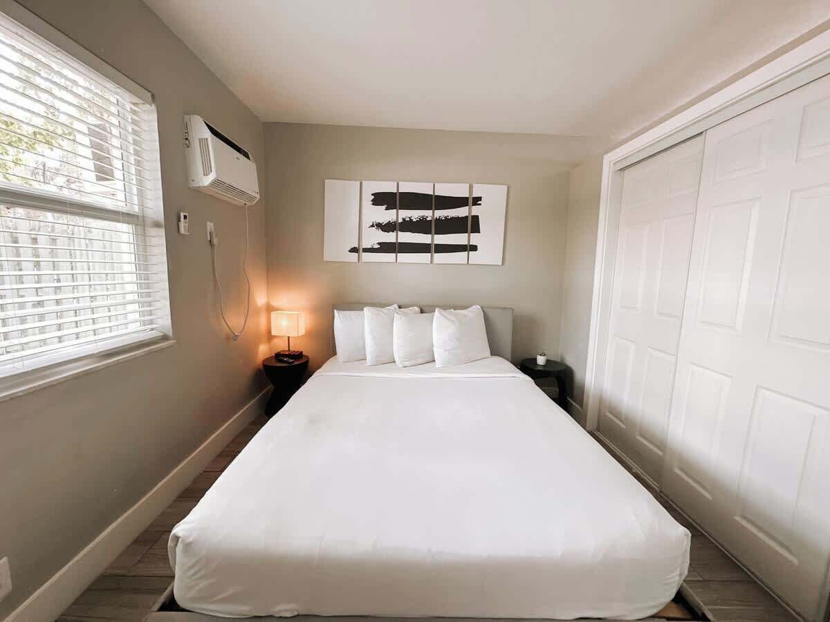 Un lit bien fait dans une pièce dotée d’une fenêtre, d’un climatiseur fixé au mur, d’une lampe de chevet et d’une porte.