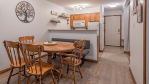Una cocina compacta con armarios de madera, un microondas y un refrigerador. Hay una mesa de comedor con sillas y un banco. Se observa un cuadro decorativo con motivos de árboles.