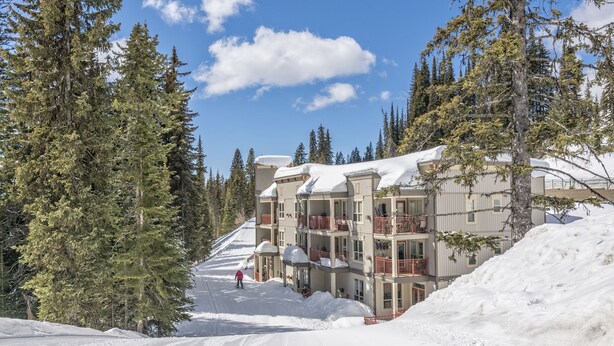 Pista de esquí nevada con un edificio que tiene balcones y un telesilla al fondo.