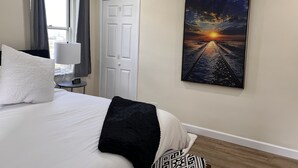 Een slaapkamer met een bed, nachtkastje, lamp, deur en een schilderij van een zonsondergang aan de muur.