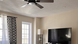 Een moderne woonkamer met een zwarte bank, een ronde salontafel, een flatscreen televisie en een plafondventilator.