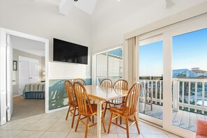 A dining area with a table and chairs, a TV mounted on the wall, and a balcony with a view of houses. - Gulf Shores Plantation-West 1338 (Gulf Shores)