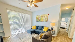 A living room with a sectional sofa, a smaller couch, a coffee table, and a floor lamp. There is a large window with blinds, a ceiling fan, and a painting on the wall. - Cozy Condo minutes away from Disney (Kissimmee)