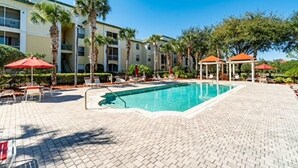 A swimming pool with lounge chairs and umbrellas, surrounded by palm trees and a multi-story building. - Cozy Condo minutes away from Disney (Kissimmee)