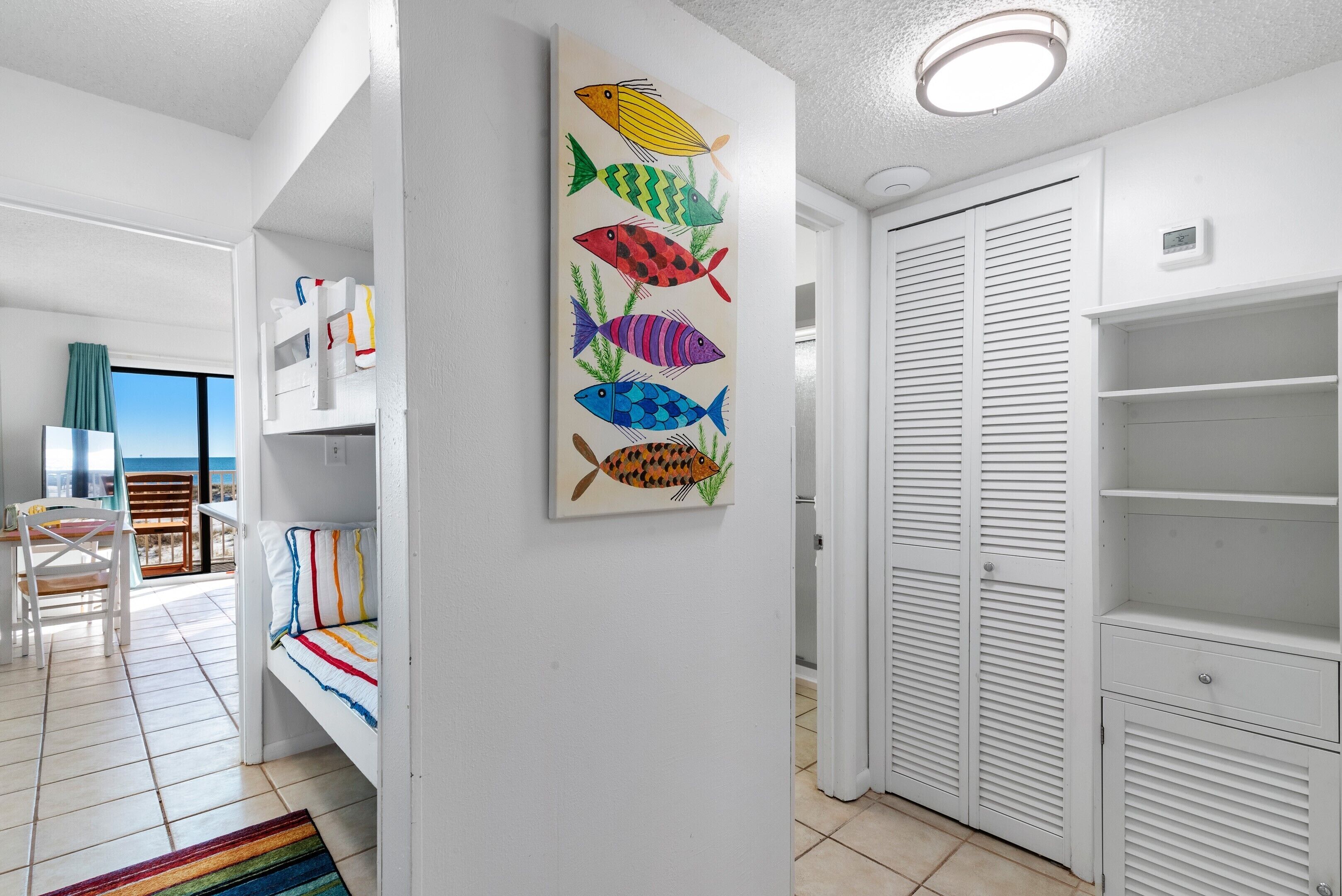 Une chambre avec un lit superposé, un tableau coloré représentant des poissons, une armoire blanche et une vue sur l’océan.