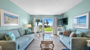 A living room with a view of a green lawn and palm trees through a glass door. - Gulf Shores Plantation-Palms 6109 (Gulf Shores)