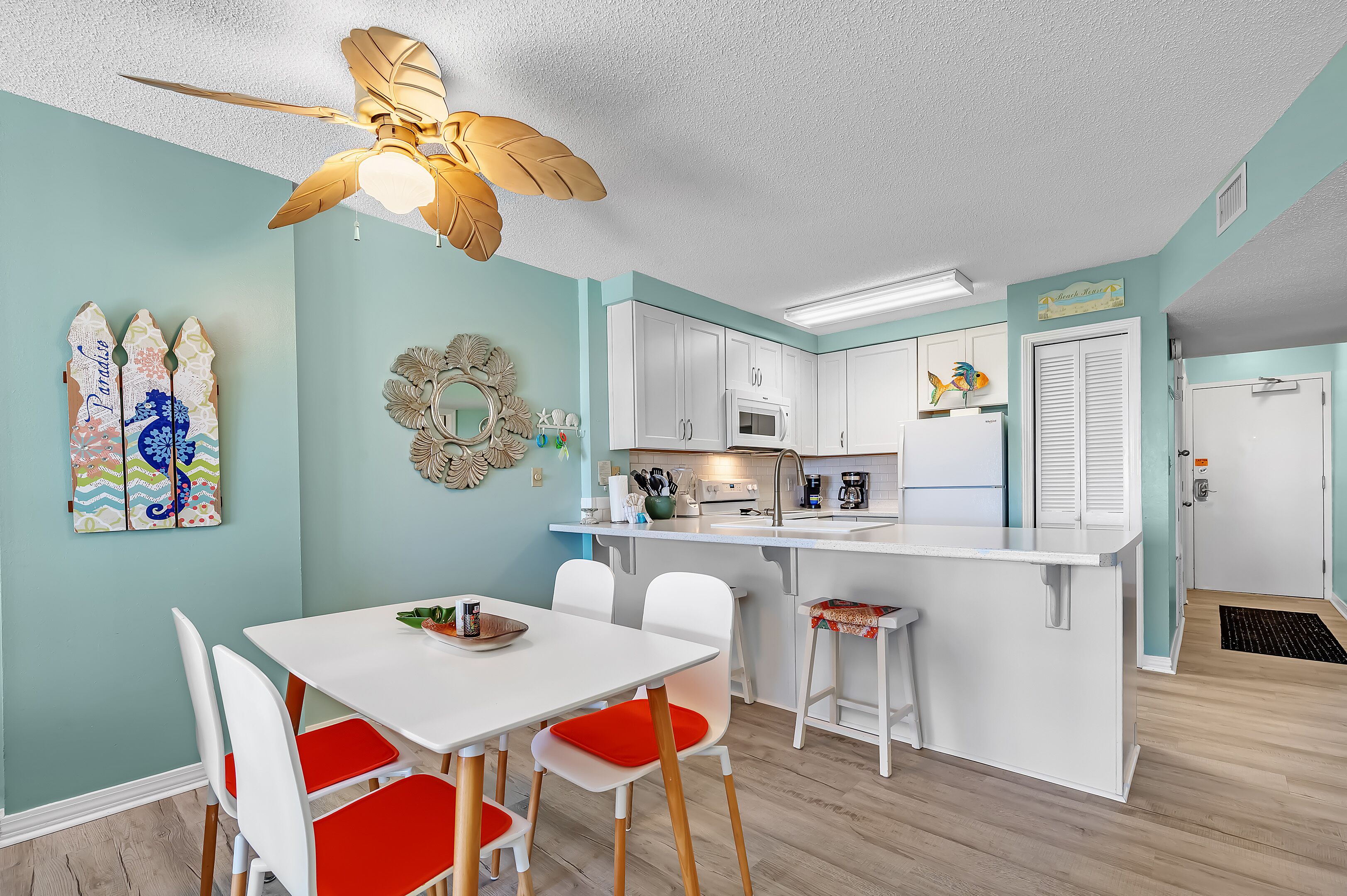 A modern kitchen with white cabinets, a microwave, and a sink. There is a dining area with a white table and red chairs. A ceiling fan is visible above the kitchen.