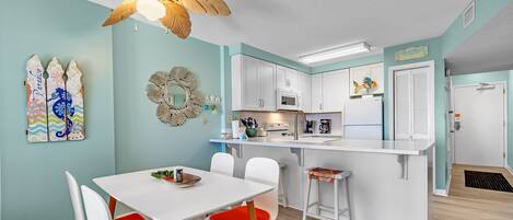 A modern kitchen with white cabinets, a microwave, and a sink. There is a dining area with a white table and red chairs. A ceiling fan is visible above the kitchen.