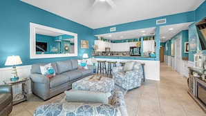 A living room with a grey sofa, patterned ottoman, and a kitchen in the background. - Gulf Shores Plantation-Dunes 5005 (Gulf Shores)