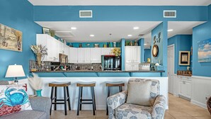 A kitchen with white cabinets, a black refrigerator, and a teal countertop. There are two bar stools and a patterned sofa in the living area. - Gulf Shores Plantation-Dunes 5005 (Gulf Shores)