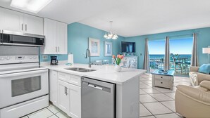 A modern kitchen with a dishwasher, microwave, and oven, a dining area with a view of the ocean, and a balcony with a table and chairs. - Gulf Shores Plantation-Palms 6406 (Gulf Shores)
