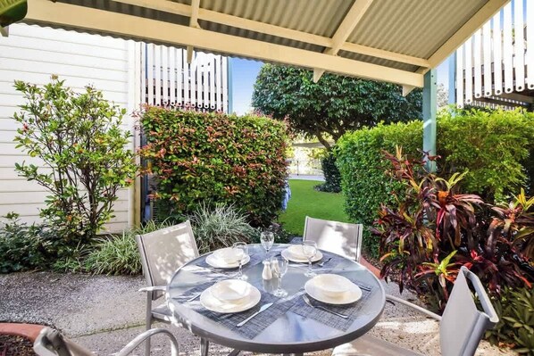 A round table set for four with chairs, positioned under a covered patio area with a white fence and greenery in the background.