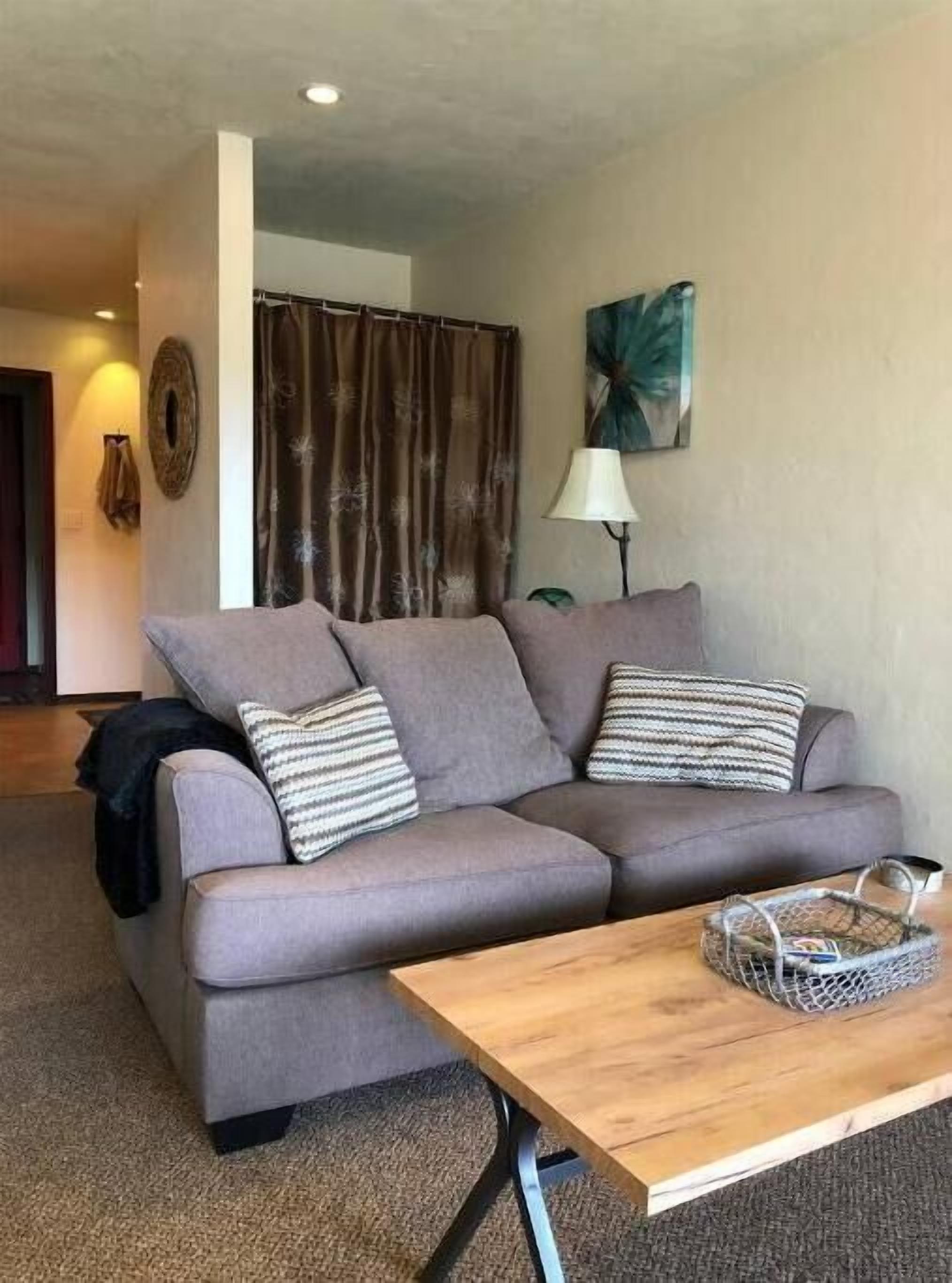 Una sala de estar con un sofá gris, dos almohadas a rayas, una mesa de centro de madera, una lámpara y un cuadro en la pared.