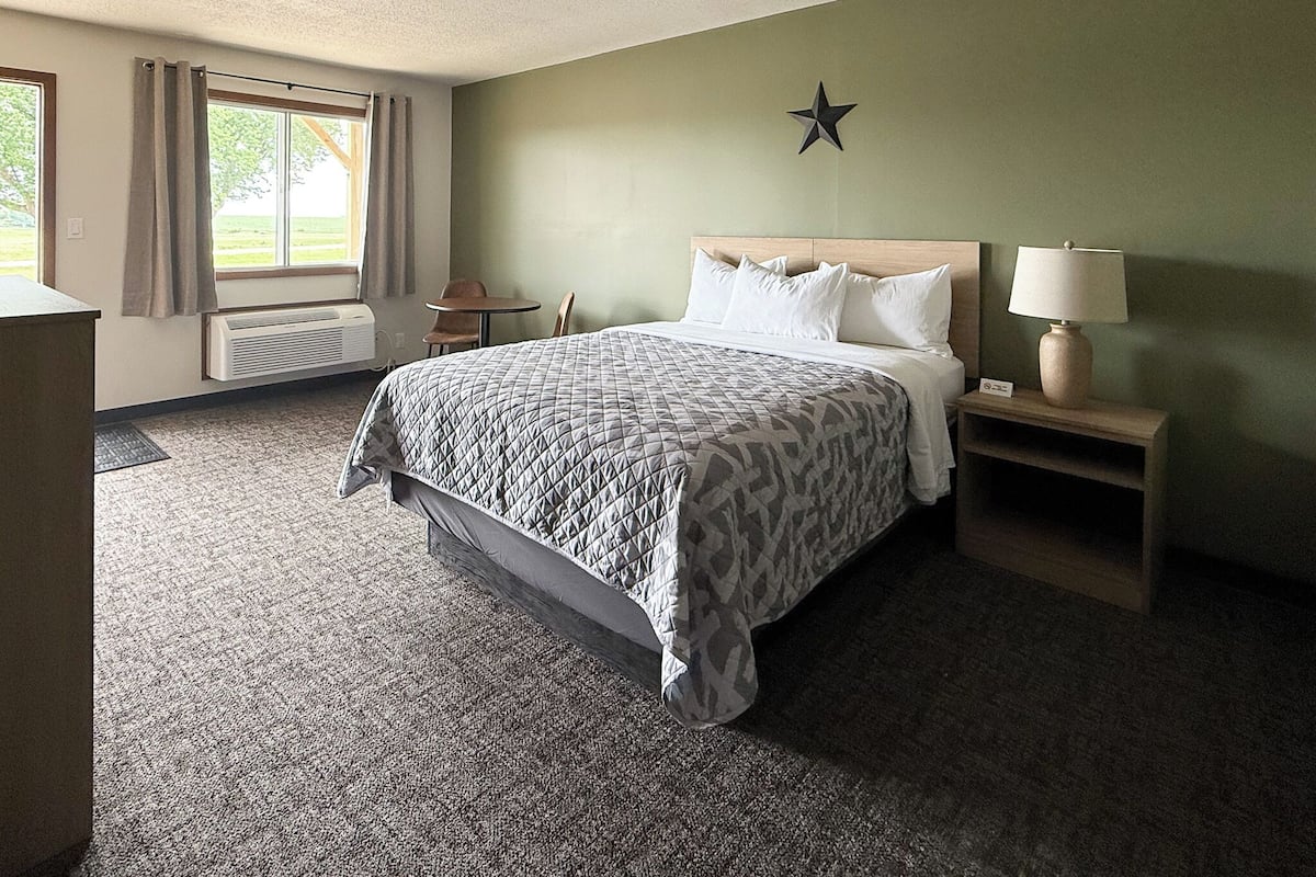 A hotel room with a bed, a nightstand with a lamp, a window with curtains, a star decoration on the wall, and a chair by the window.