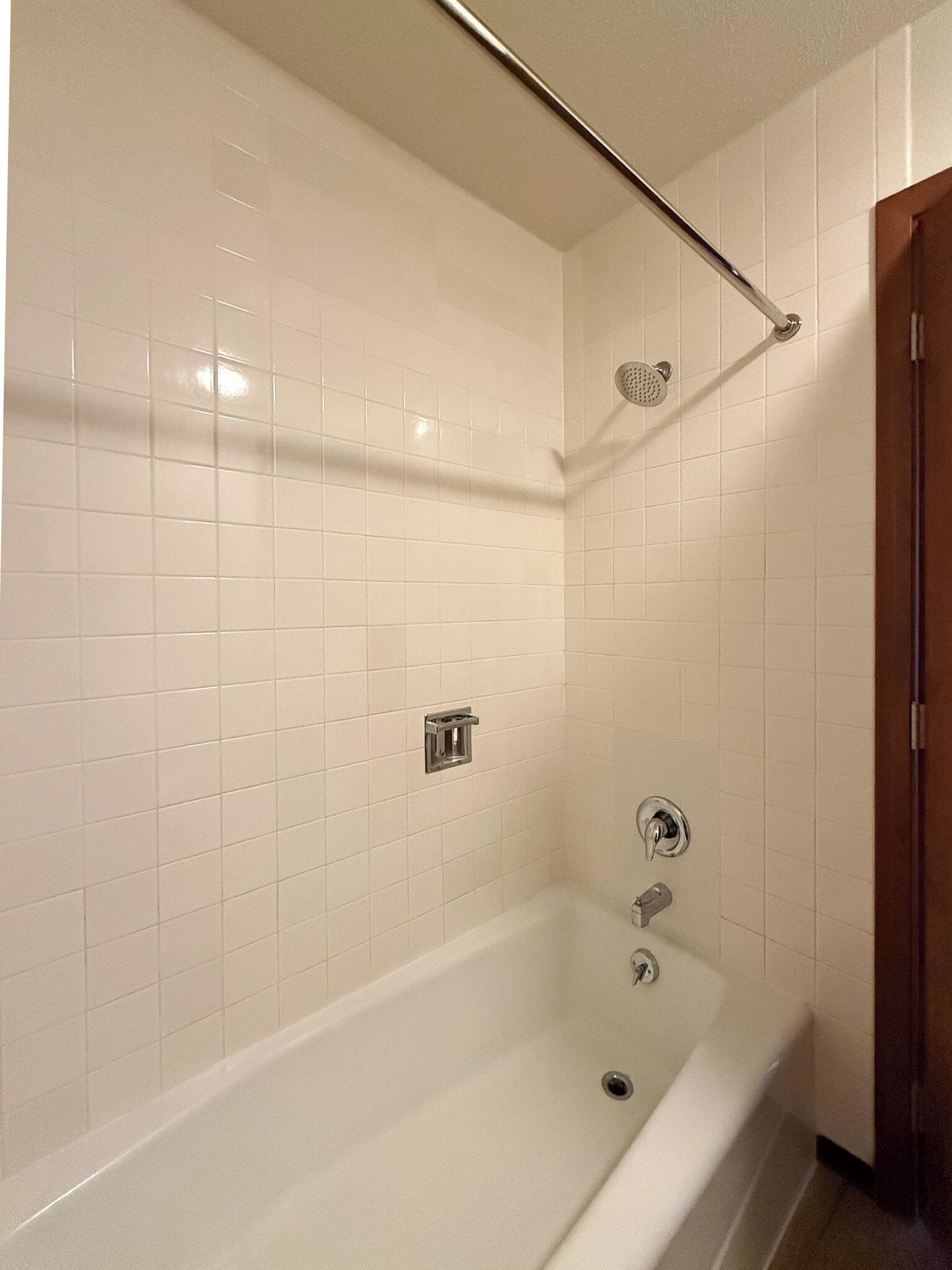 Un baño con paredes de azulejos blancos, una bañera, un cabezal de ducha y un perchero para toallas.