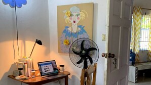 Una habitación con un escritorio de madera, una computadora portátil, un ventilador, una silla, un cuadro y una puerta que da a otra habitación.