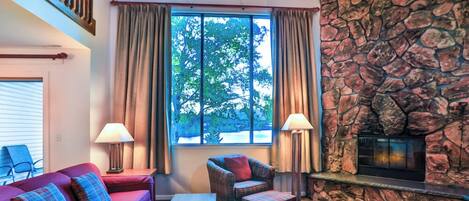 A cozy living room with a stone fireplace, a red sofa, plaid cushions, and a view of trees through large windows.