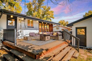 A covered outdoor seating area with a fire pit, wooden deck, and a staircase leading to a door.