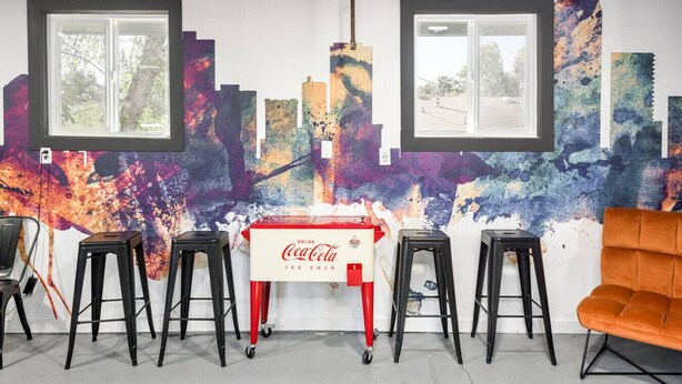 A room with a mural, two windows, a Coca-Cola refrigerator, and four stools.