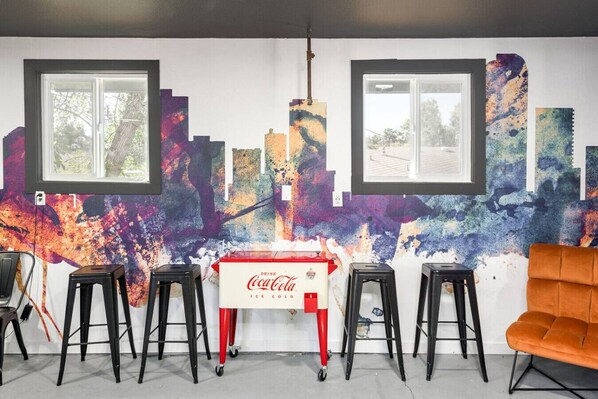 A room with a mural, two windows, a Coca-Cola refrigerator, and four stools.