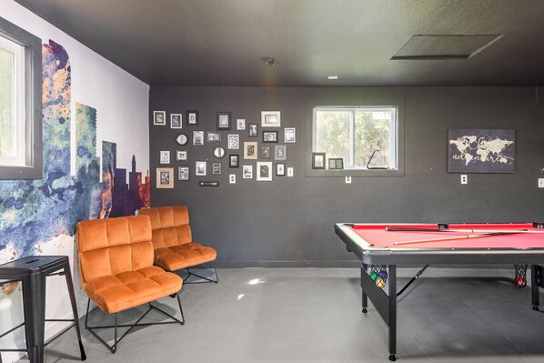 A room with a pool table, two orange chairs, and a wall decorated with framed pictures.