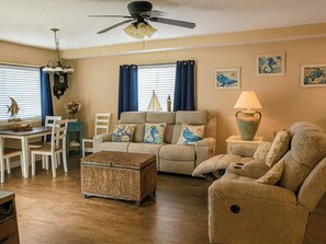 Un salon comprenant un canapé, des fauteuils, une table basse, une table à manger avec des chaises, un ventilateur de plafond et des œuvres d’art accrochées aux murs.