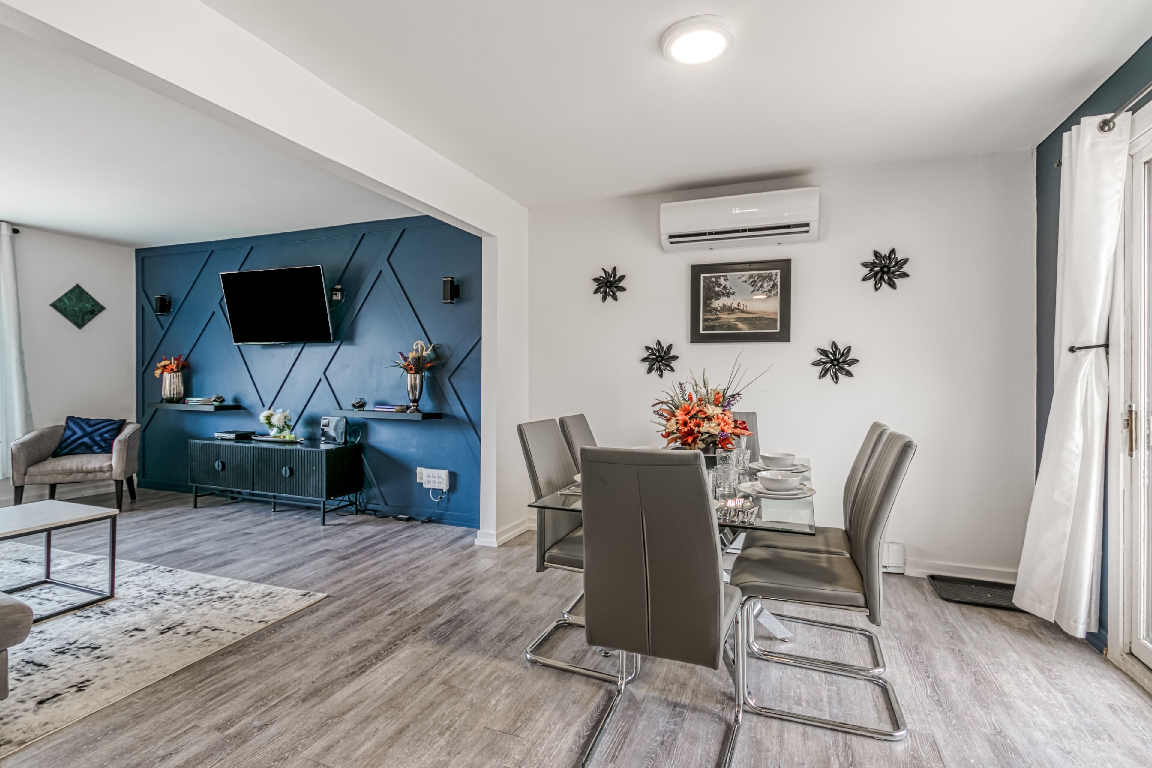 A modern living room with a blue accent wall, a mounted TV, a dining area with a table and chairs, and a decorative wall with star-shaped ornaments.