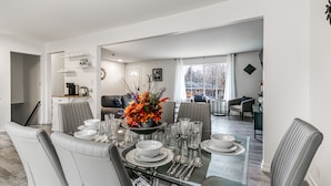 Un espace repas moderne avec une table en verre disposée pour six personnes, entourée de chaises à revêtement gris. Un vase décoratif contenant des fleurs orange est placé au centre.