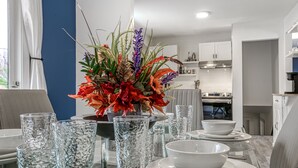 Un coin repas avec une table dressée pour quatre personnes, un vase de fleurs et, au fond, une cuisine moderne.