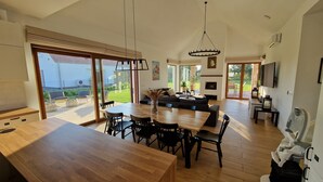 Un vaste espace de vie comprenant une table à manger en bois, des chaises noires, un canapé et un téléviseur. Une porte-fenêtre coulissante donne accès à un patio aménagé avec du mobilier d’extérieur.