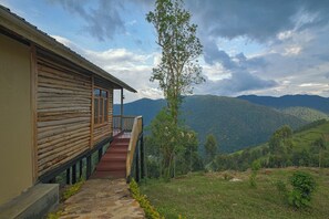 A wooden cabin with a stone pathway leading to a wooden deck, overlooking a mountainous landscape. - Orugano Bwindi Lodge (Nkuringo)