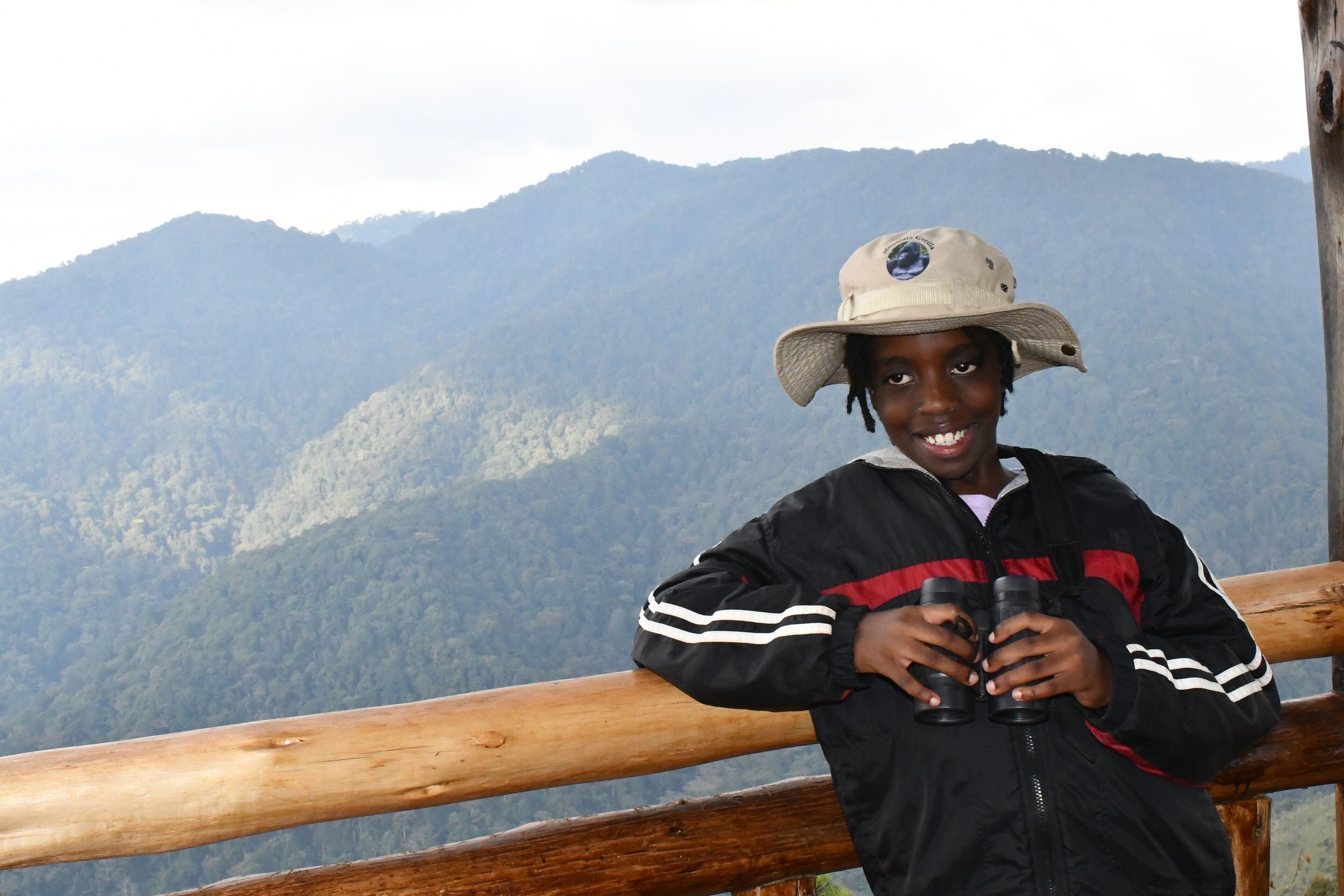 A person holding binoculars, standing on a wooden platform with a mountainous landscape in the background.