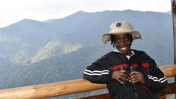 A person holding binoculars, standing on a wooden platform with a mountainous landscape in the background.