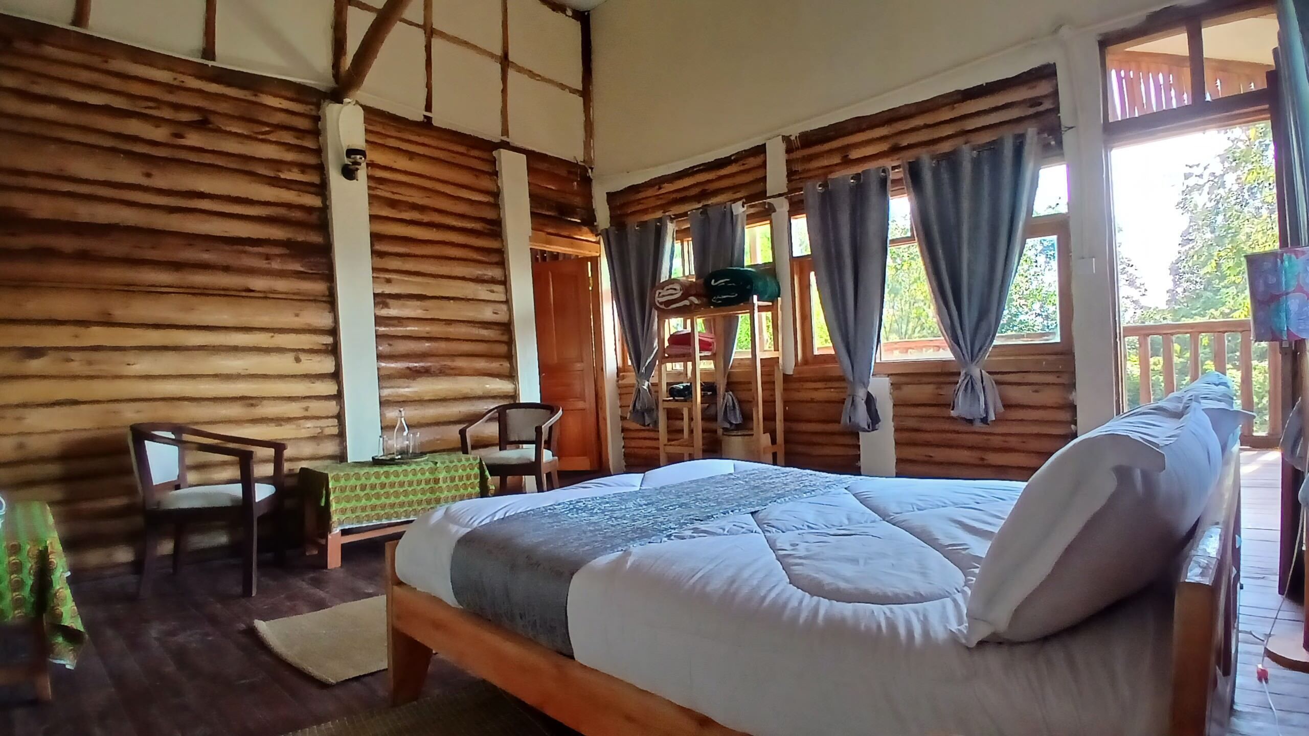 A log cabin room with a bed, a chair, a table, and a window with curtains.
