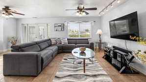 A modern living room with a gray sectional sofa, a glass coffee table, a flat-screen TV, and a ceiling fan. - Gulf Coast Get-Away! Heated pool, sleeps 10! 69th (Largo)